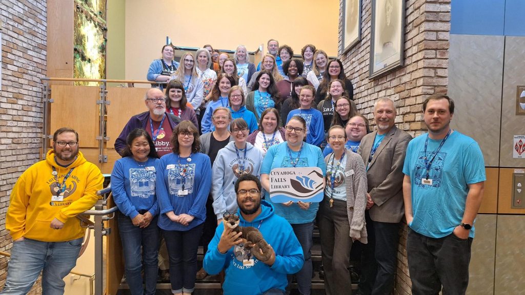 A group photo of the Cuyahoga Falls Library staff standing in rows on the library's stairs, wearing library tshirts.