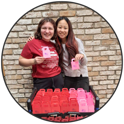 Two teen girls holding a small shopping bag papercraft in front of a cart full of those small bags.