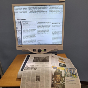 A magnifying device that is aimed at a newspaper and magnifying its text onto a monitor with adjustable dials.